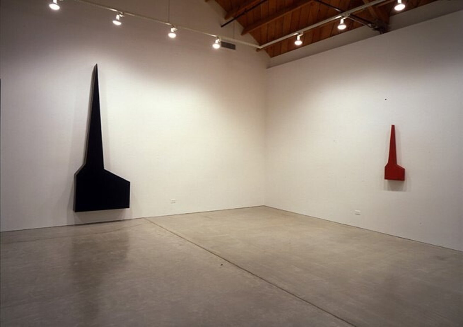 A minimalist sculpture featuring a sharp red spire rising from a flat base against a plain wall.