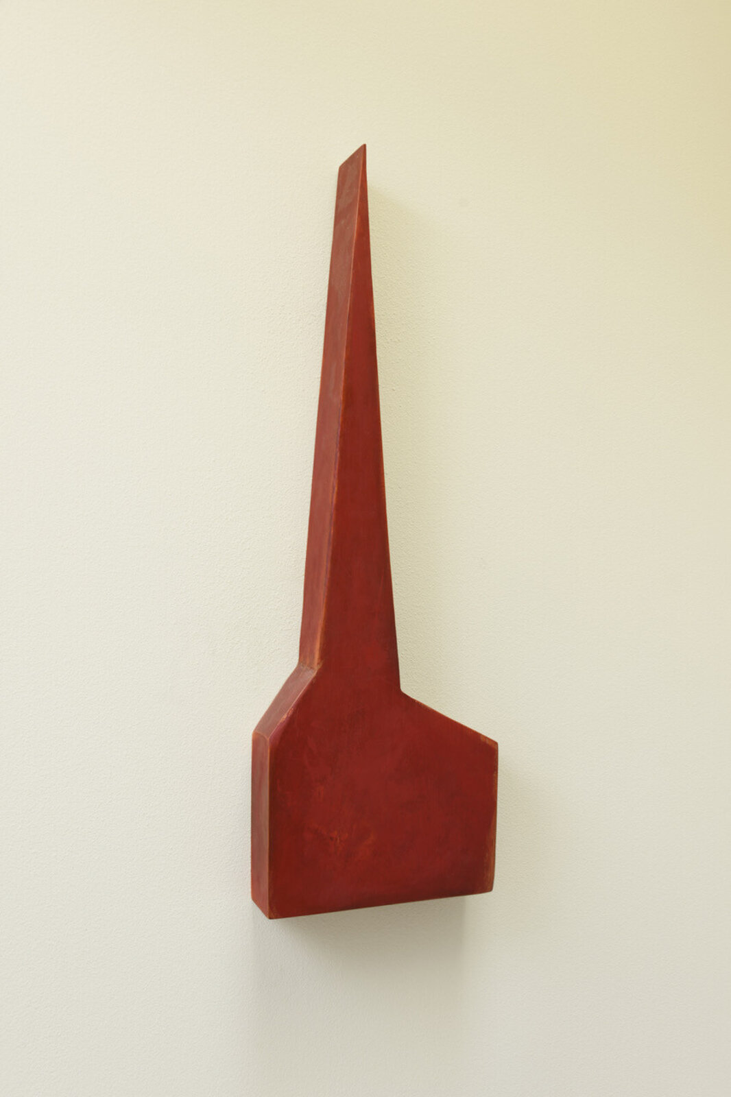 A minimalist sculpture featuring a sharp red spire rising from a flat base against a plain wall.