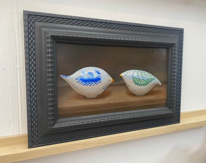 A framed diorama featuring two ceramic bird figures facing each other.