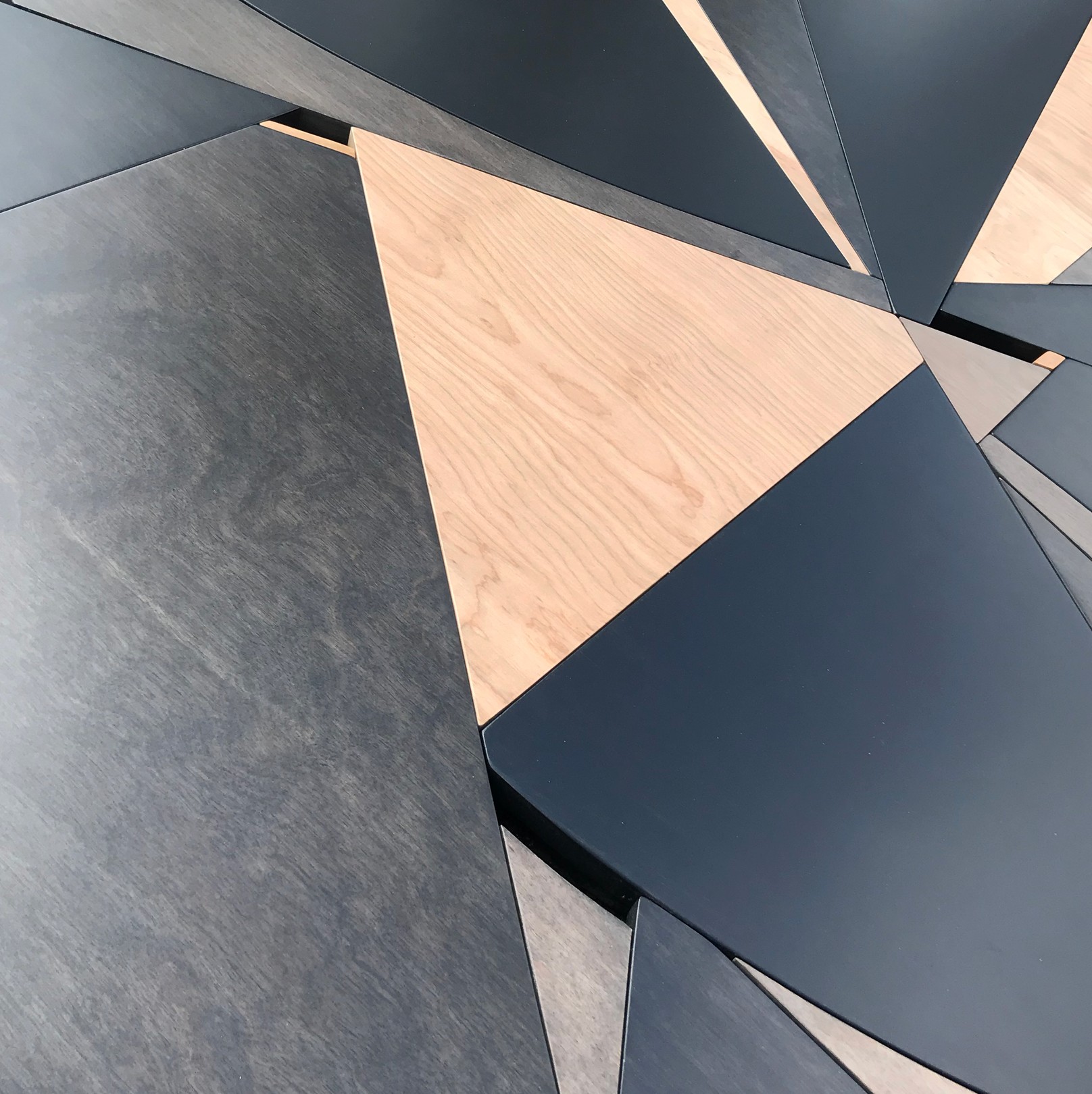 A geometric wooden sculpture composed of layered triangular panels in black, light wood, and dark wood resting on asphalt.
