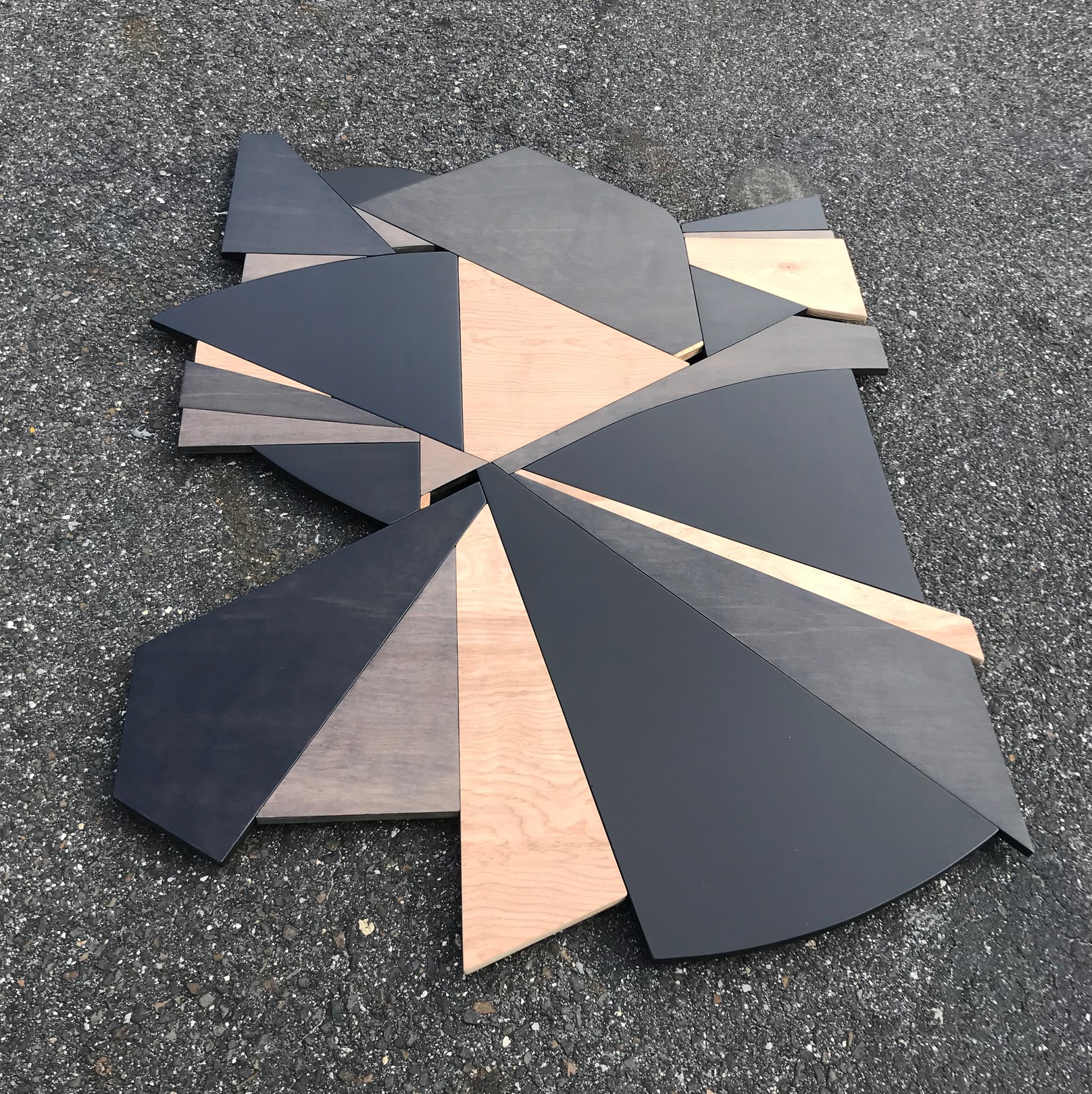 A geometric wooden sculpture composed of layered triangular panels in black, light wood, and dark wood resting on asphalt.