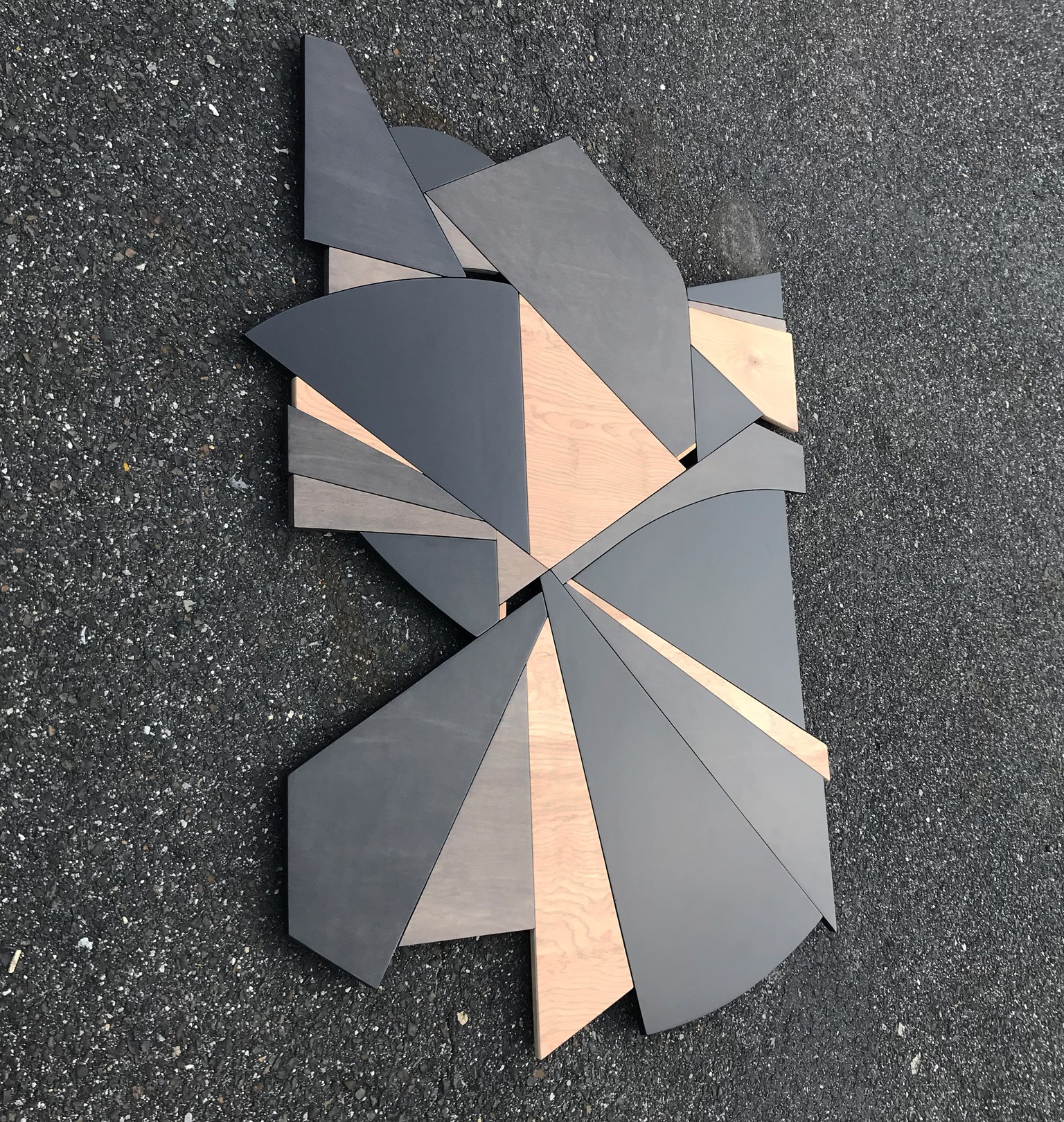 A geometric wooden sculpture composed of layered triangular panels in black, light wood, and dark wood resting on asphalt.
