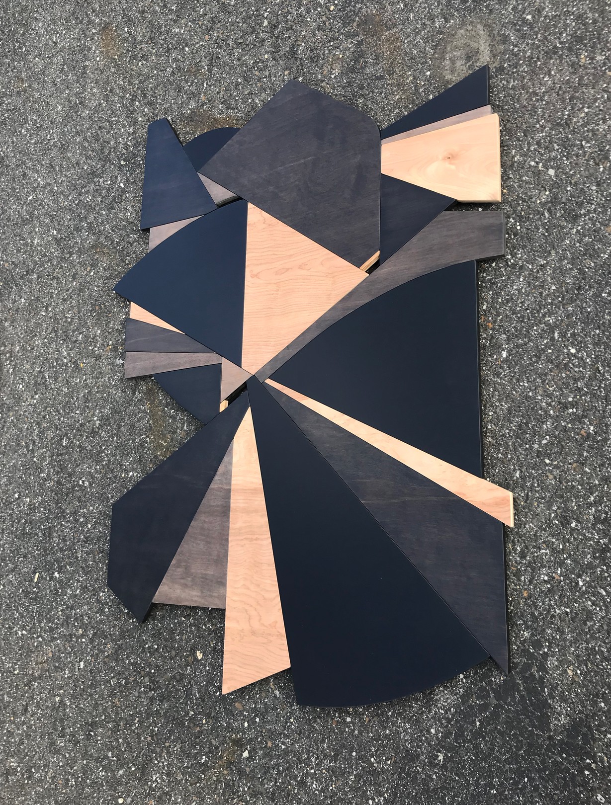 A geometric wooden sculpture composed of layered triangular panels in black, light wood, and dark wood resting on asphalt.