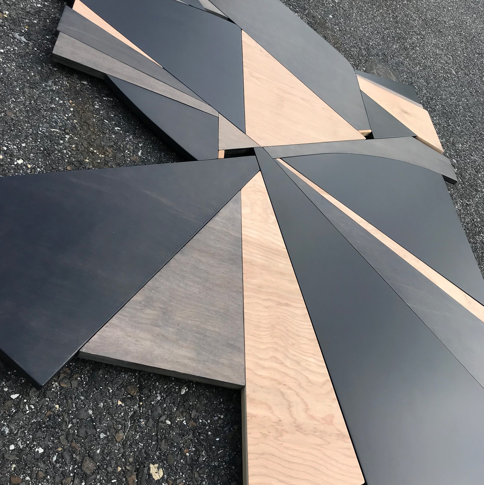 A geometric wooden sculpture composed of layered triangular panels in black, light wood, and dark wood resting on asphalt.