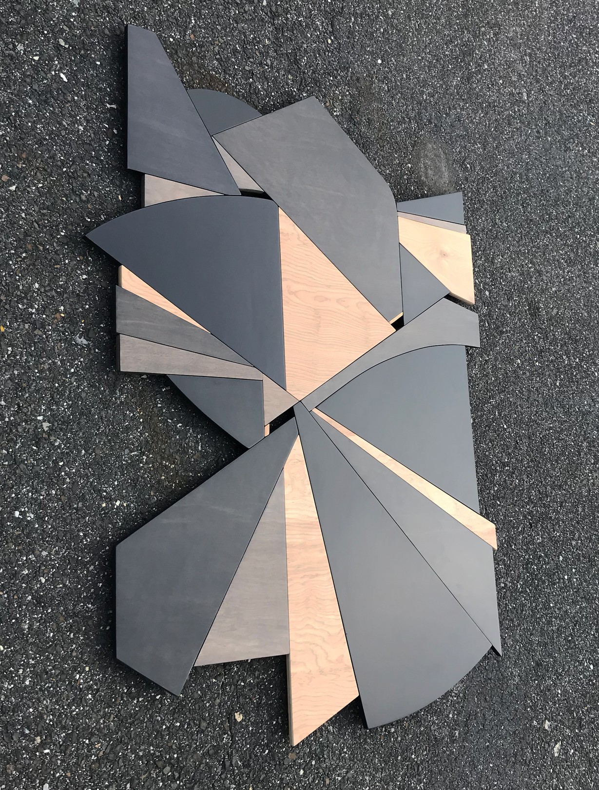 A geometric wooden sculpture composed of layered triangular panels in black, light wood, and dark wood resting on asphalt.