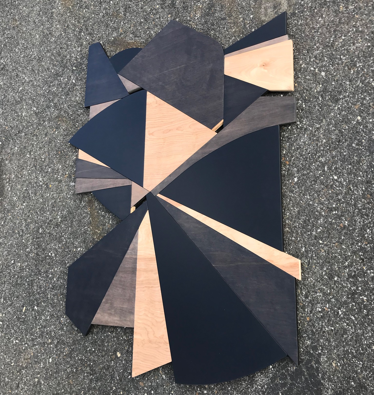 A geometric wooden sculpture composed of layered triangular panels in black, light wood, and dark wood resting on asphalt.