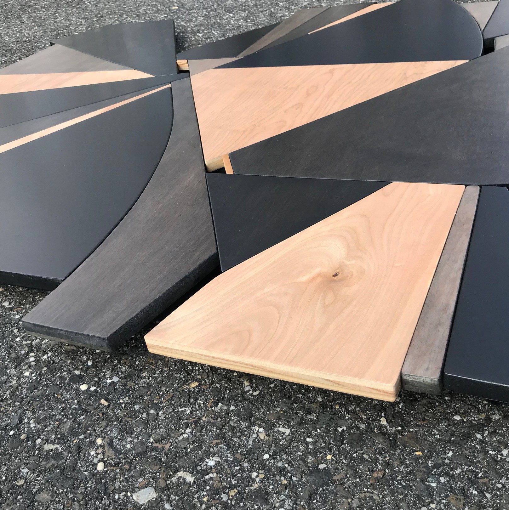 A geometric wooden sculpture composed of layered triangular panels in black, light wood, and dark wood resting on asphalt.