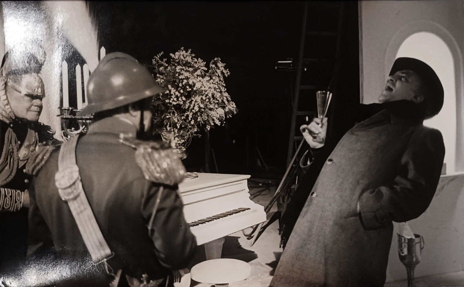 A dramatic black-and-white scene featuring a soldier, a figure in costume, and another person celebrating with champagne.