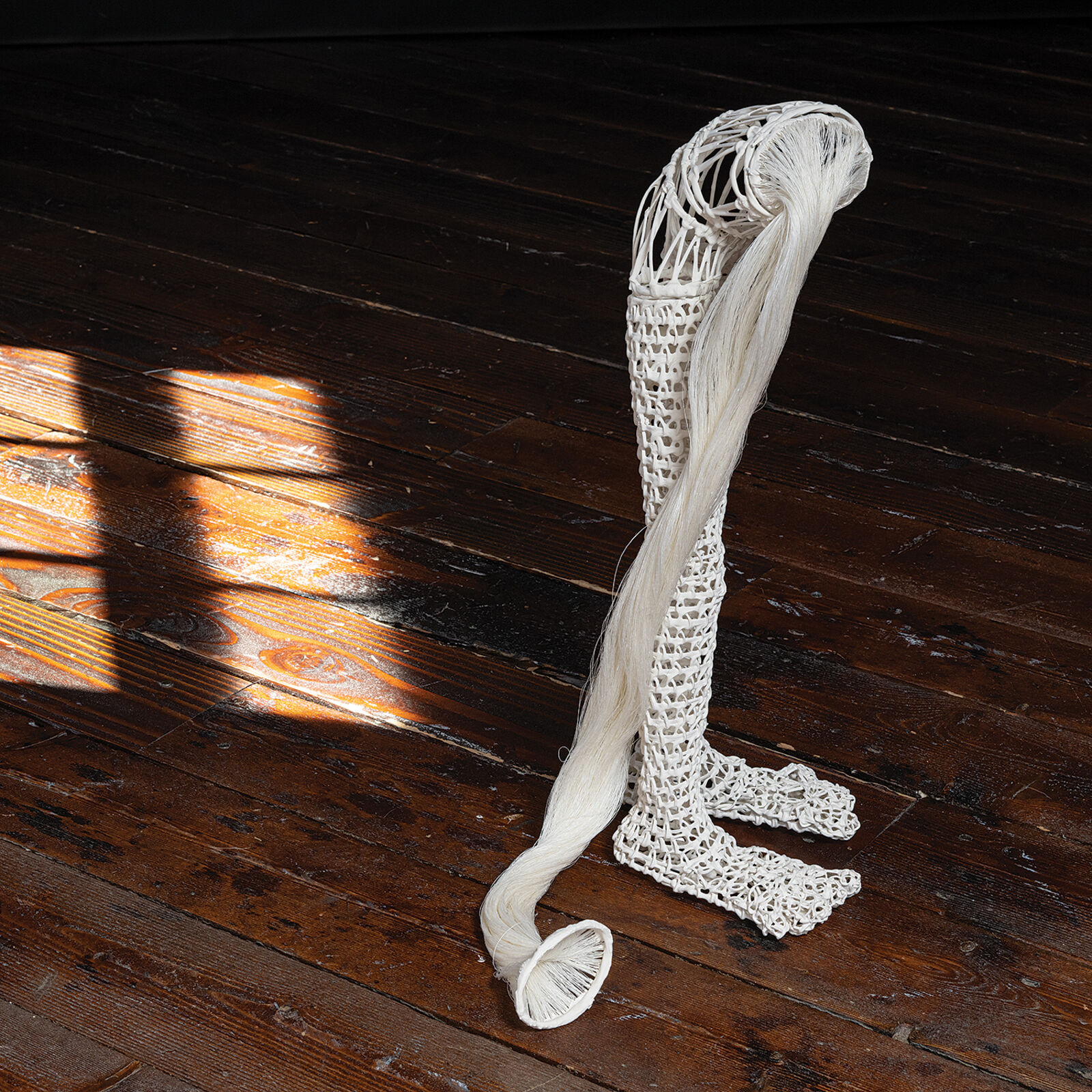 A white, crocheted sculpture resembling a stylized leg or boot standing on a dark wooden floor.