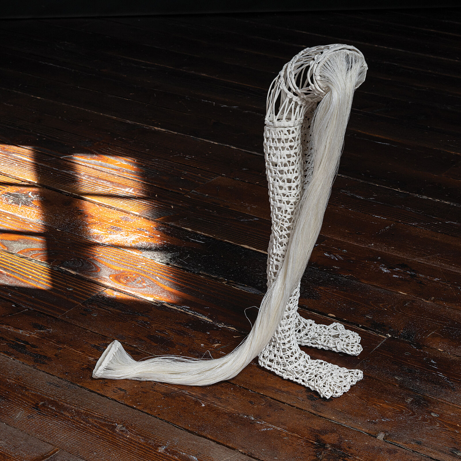 A white, crocheted sculpture resembling a stylized leg or boot standing on a dark wooden floor.