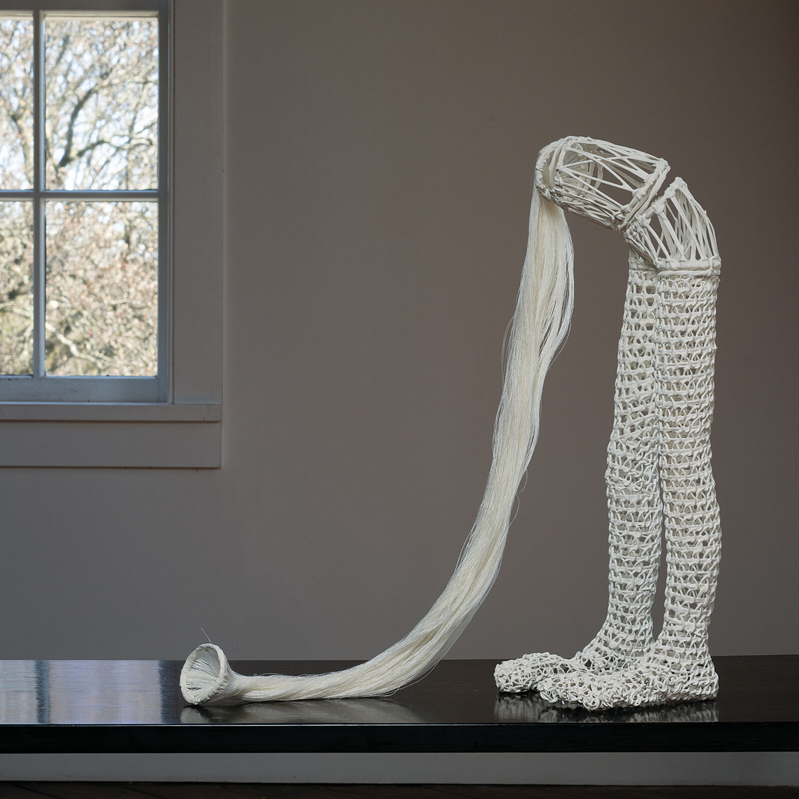 A white, crocheted sculpture resembling a stylized leg or boot standing on a dark wooden floor.