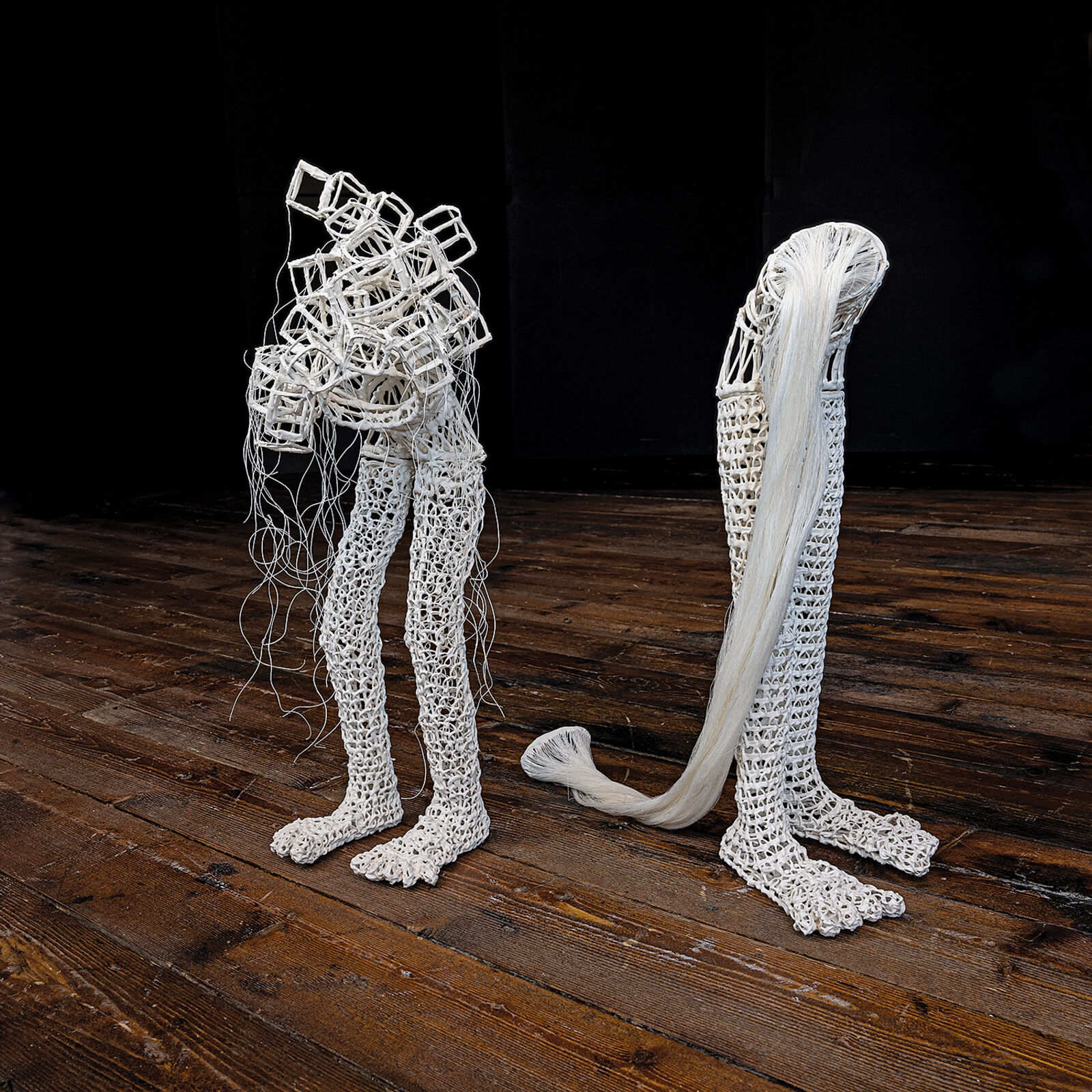 A white, crocheted sculpture resembling a stylized leg or boot standing on a dark wooden floor.