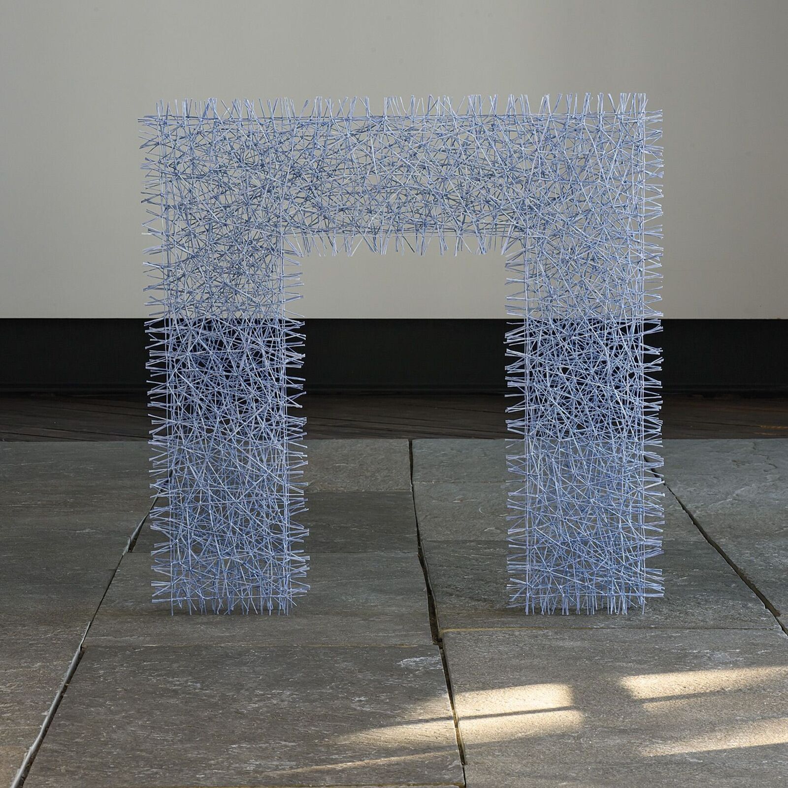 A light blue wire sculpture shaped like a stylized letter 'H' stands on a stone floor.