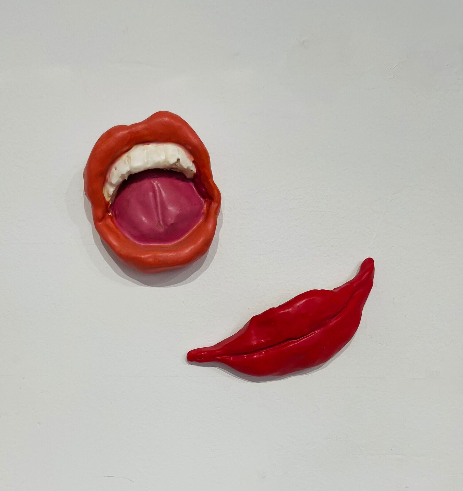 A sculptural composition featuring a mouth with teeth and tongue, alongside a detached red lip, set against a plain white background.