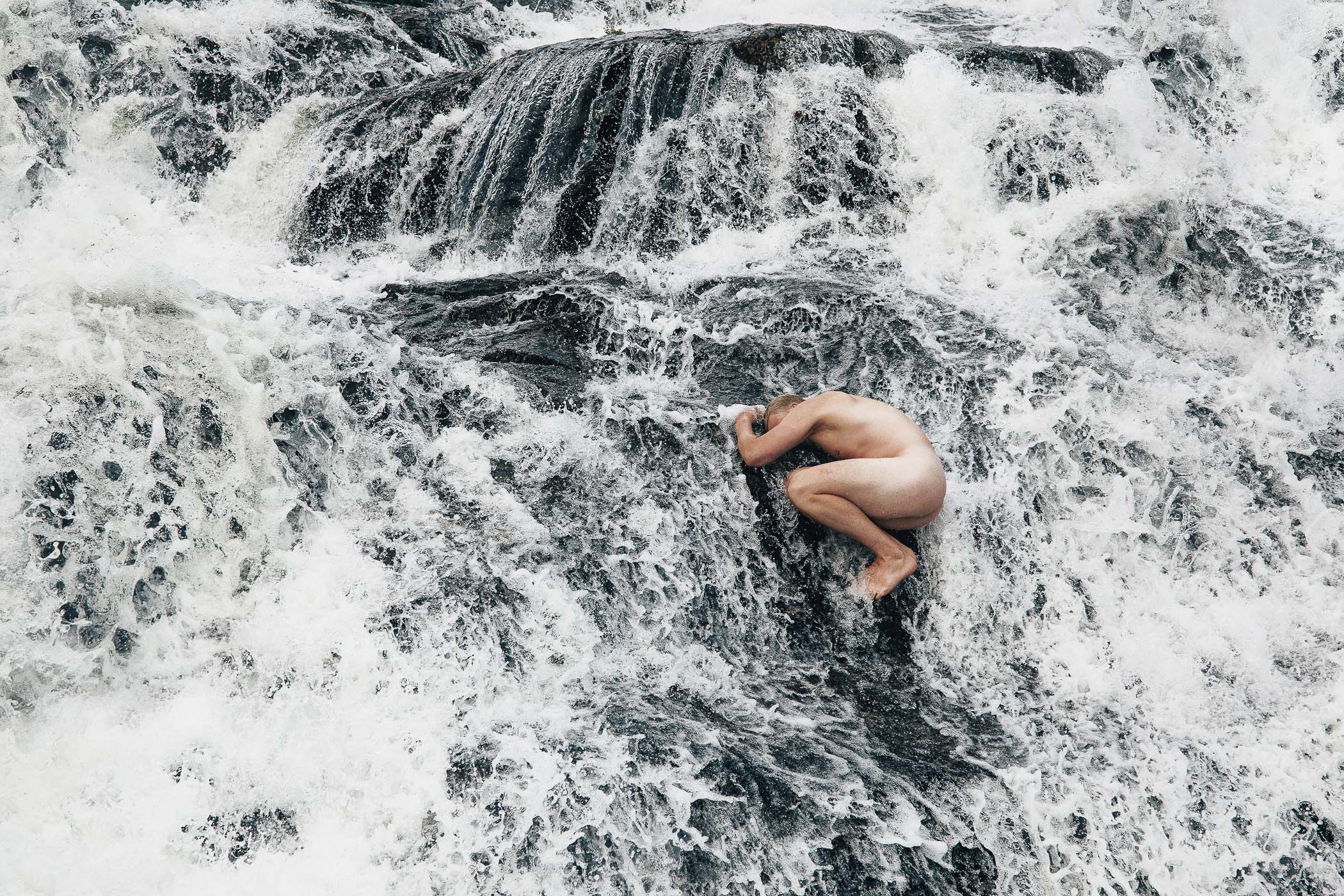 A naked man sitting on a rock in the middle of a waterfall.