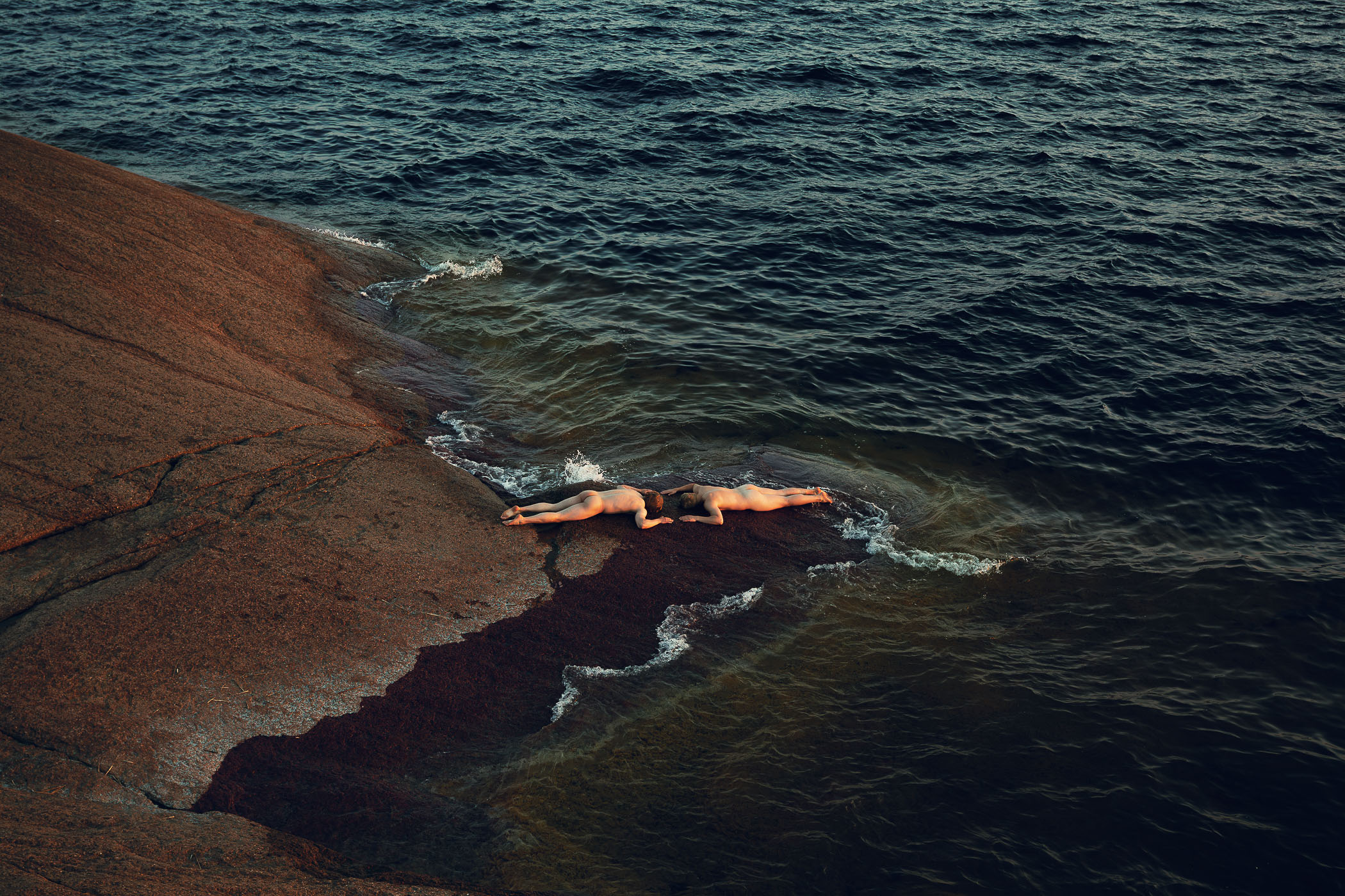 A couple of people laying on top of a rock next to the ocean.