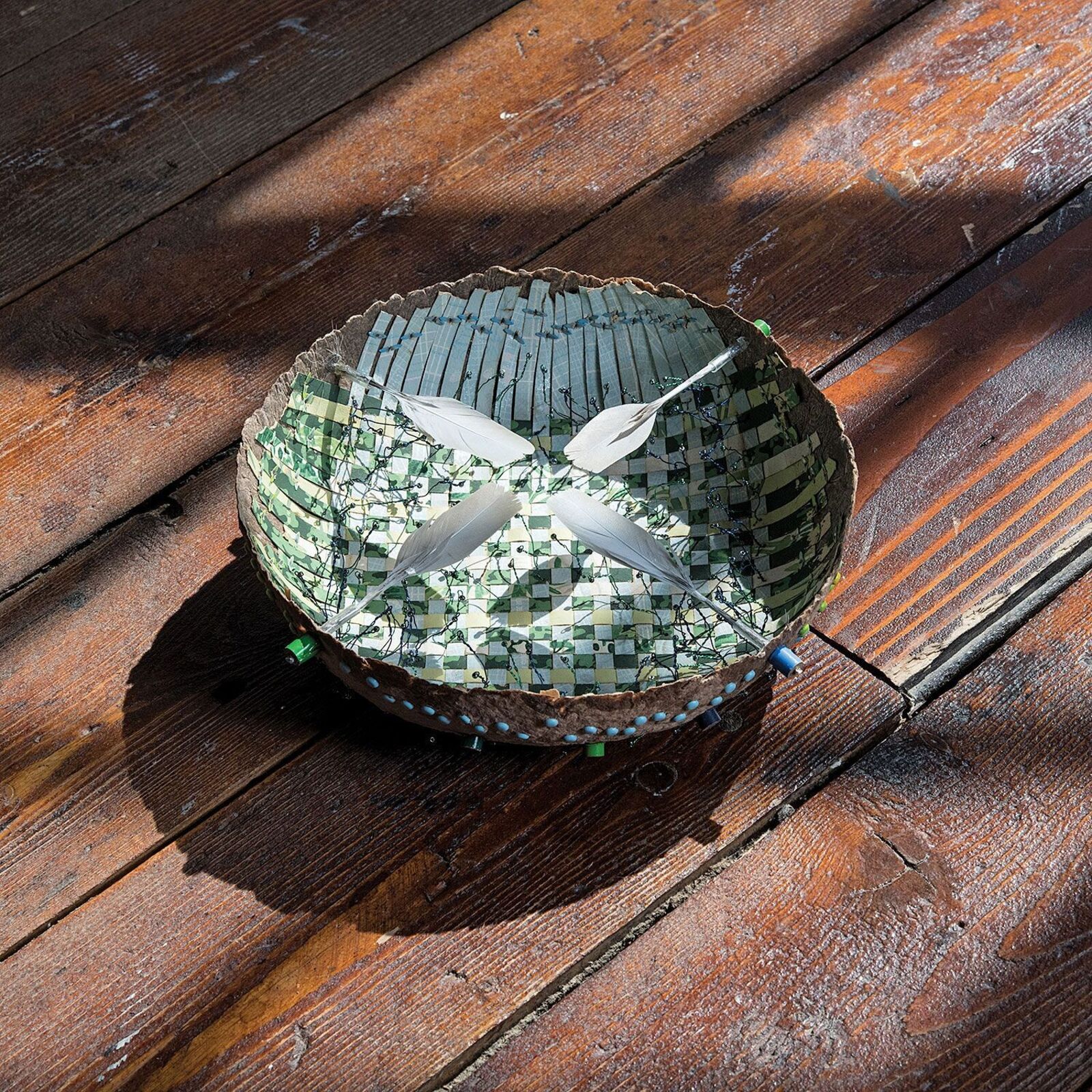 A handcrafted bowl with a mosaic interior and feathers rests on weathered wooden planks.