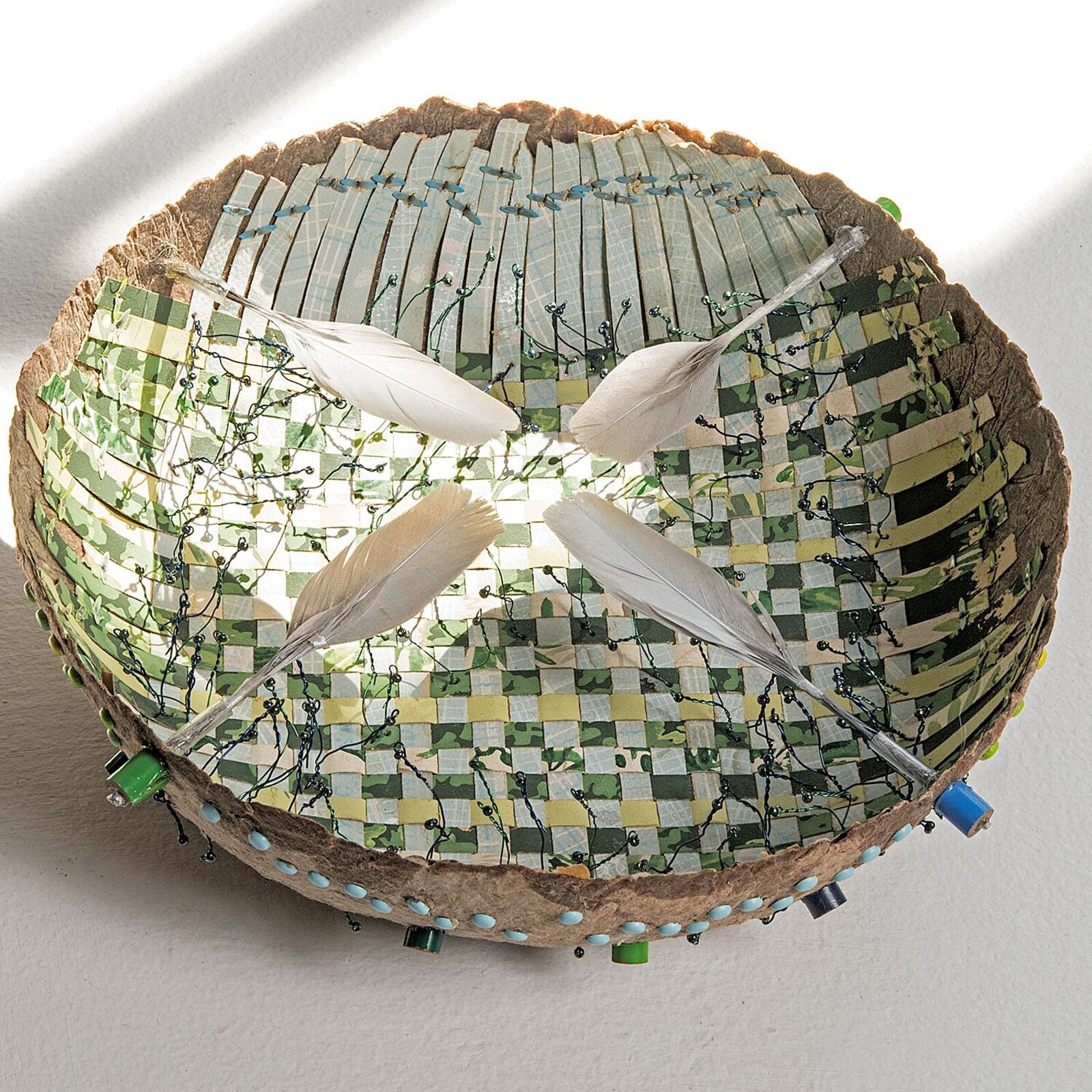 A handcrafted bowl with a mosaic interior and feathers rests on weathered wooden planks.
