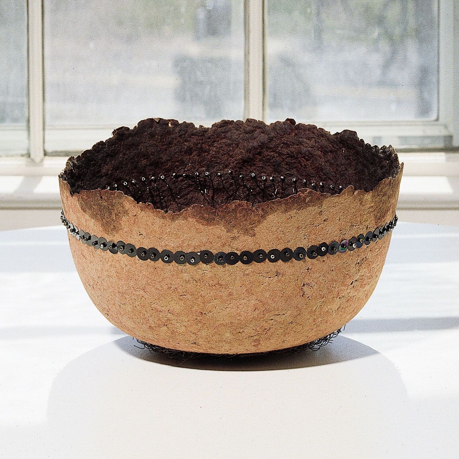 A textured bowl filled with dark soil, adorned with black sequins and wire.
