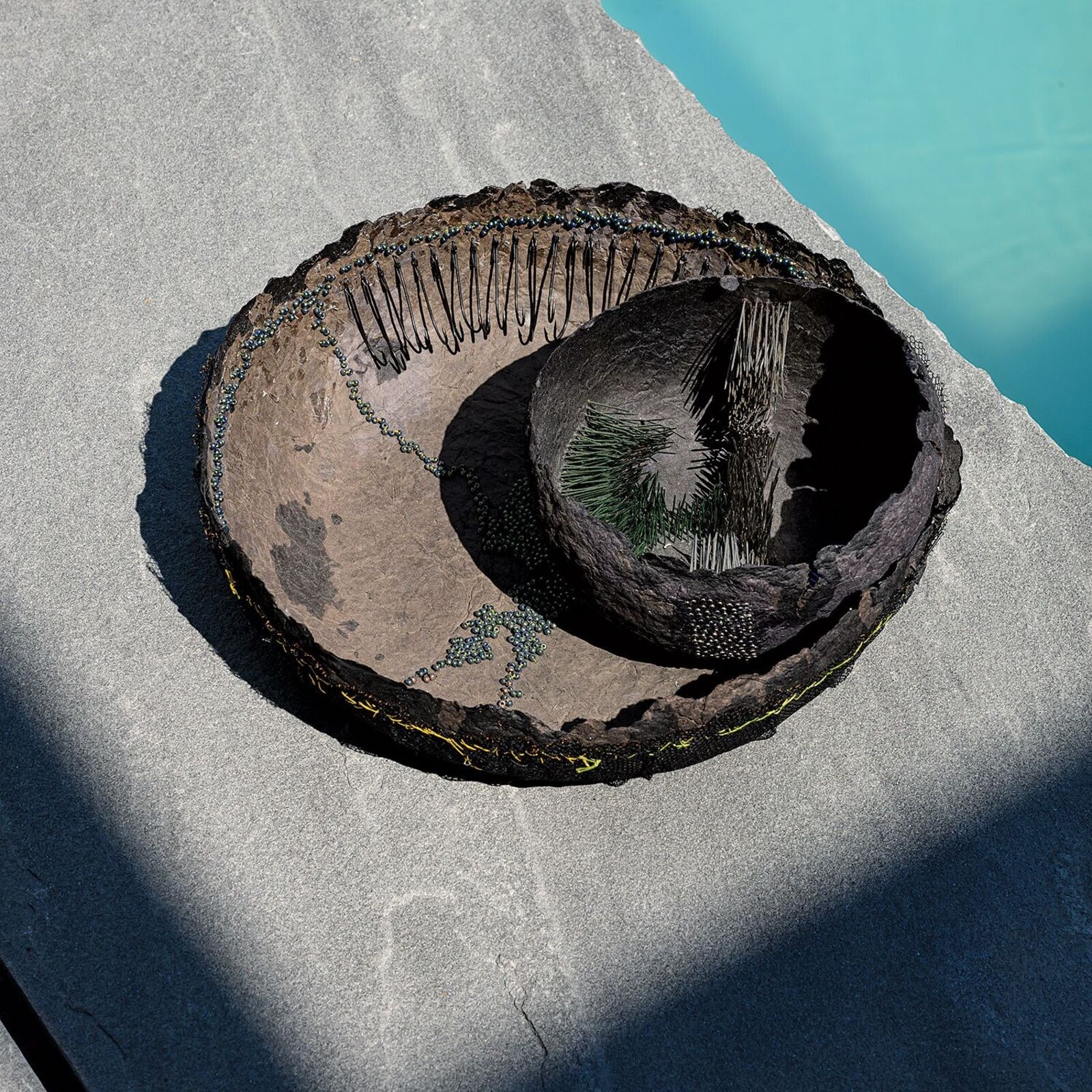 A high-angle view of a textured, bowl-like sculpture resting on a stone surface next to water.