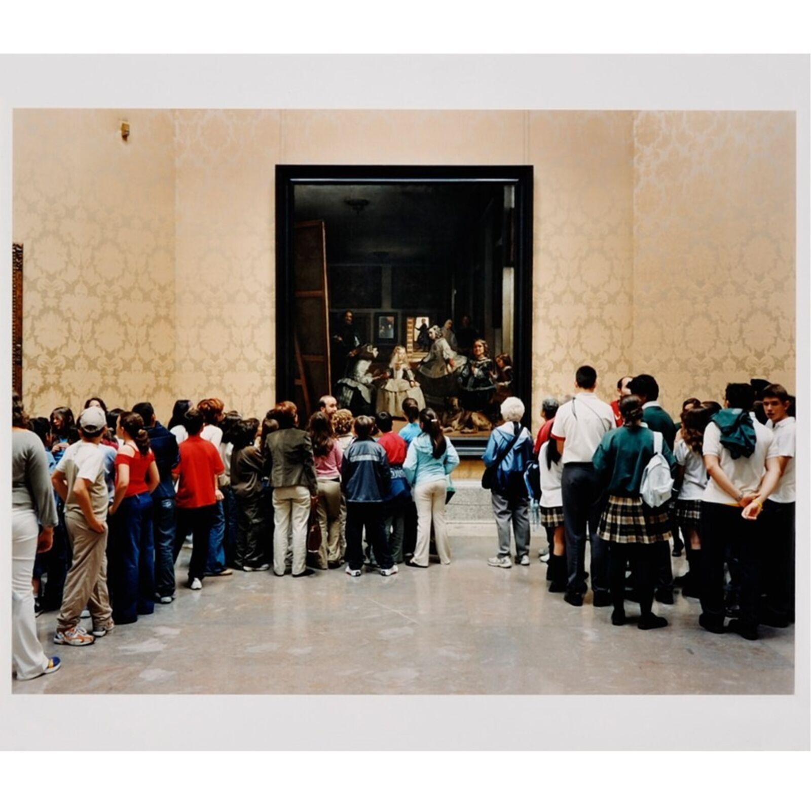 A group of people in a museum hall look at a large painting displayed behind glass.