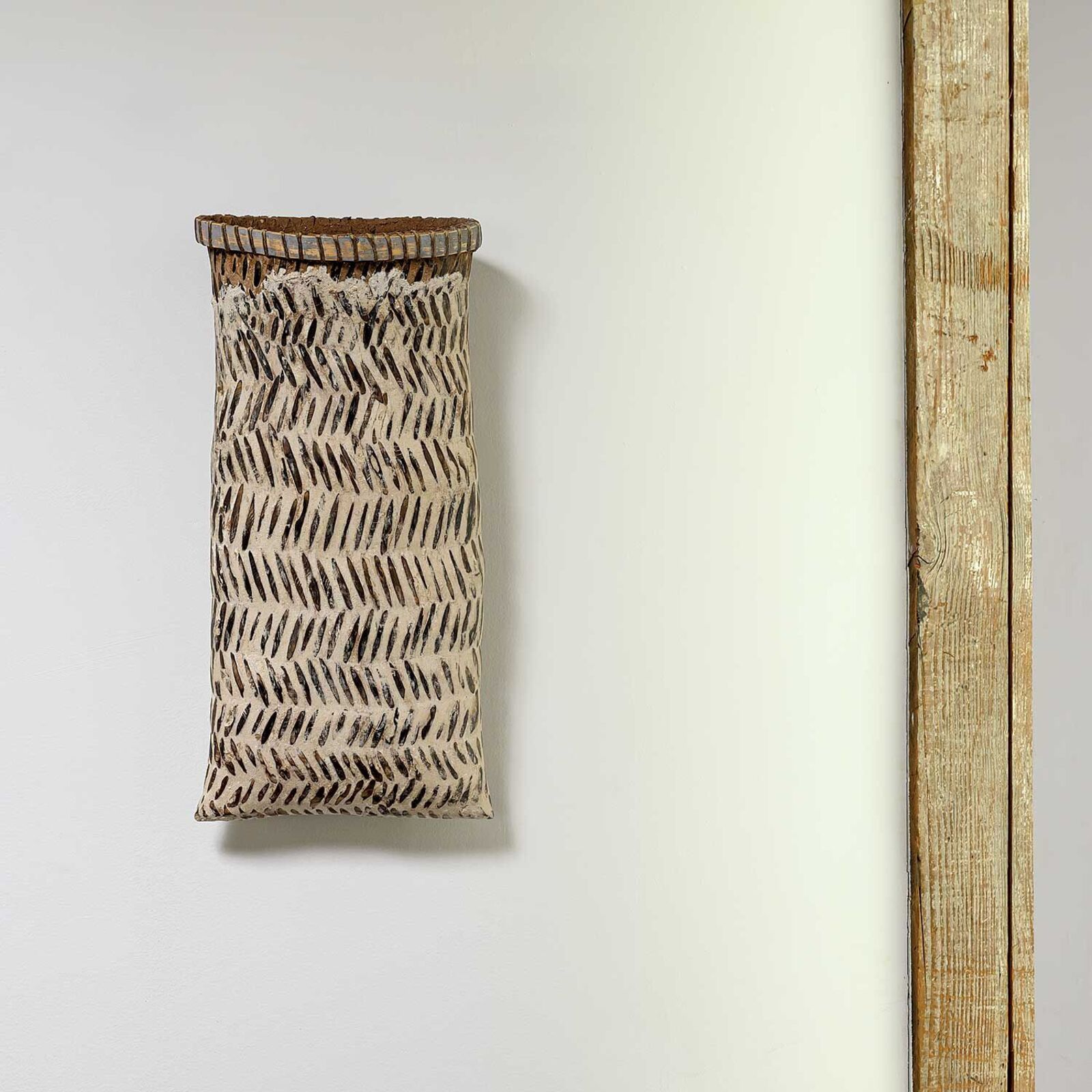 A vertically oriented woven basket with a textured pattern hangs against a plain wall next to a wooden beam.