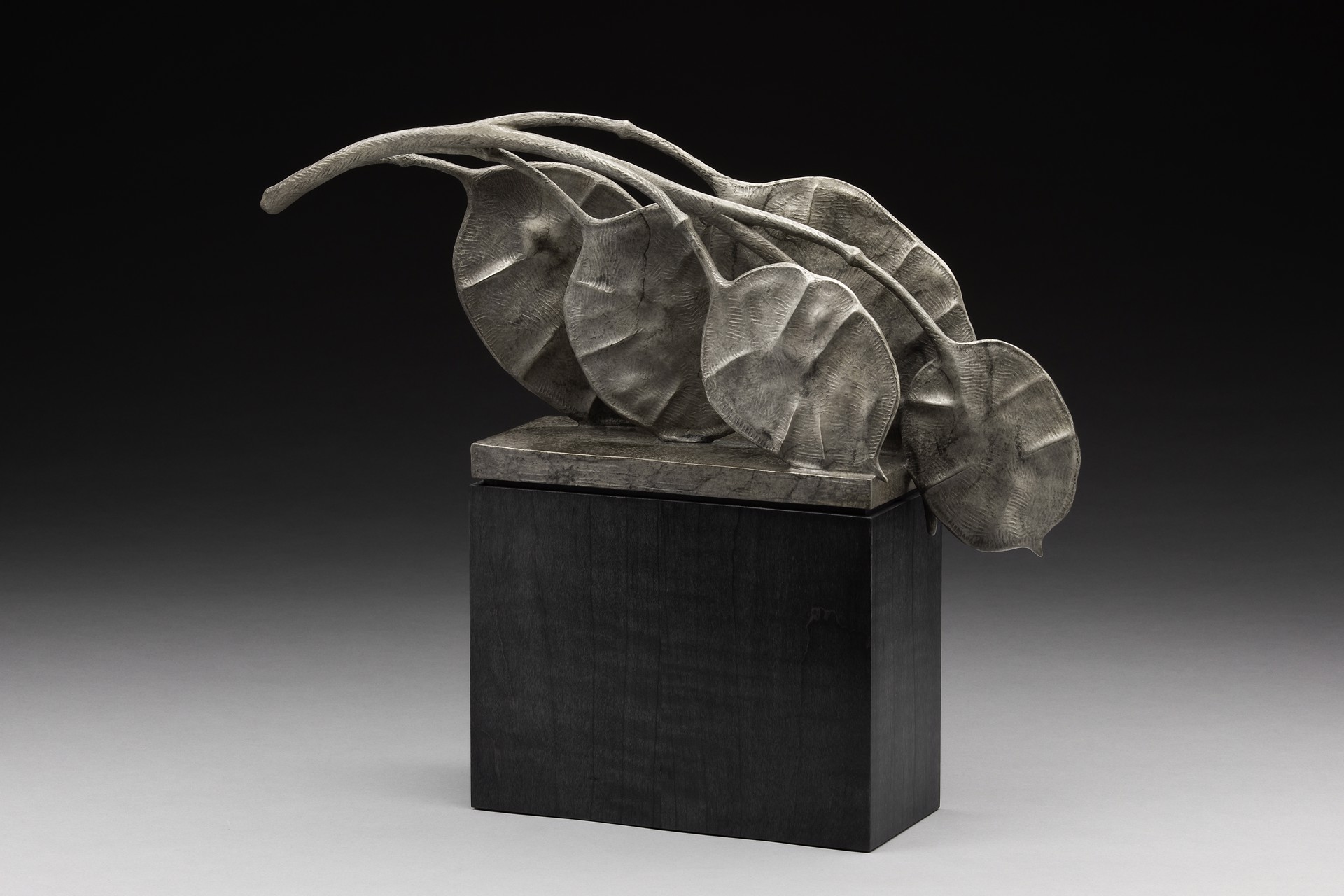 A sculpture of leaves on a black pedestal on a gray background.
