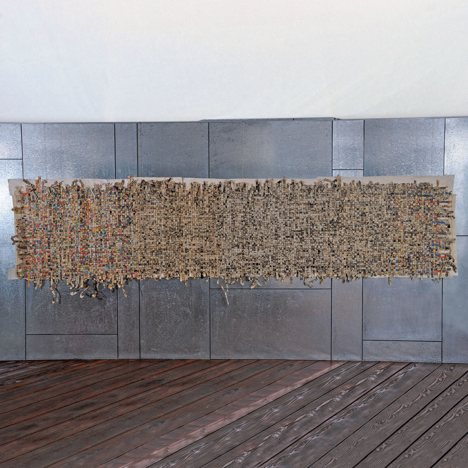 A rectangular textile artwork with a dense, textured surface displayed against a metallic wall.