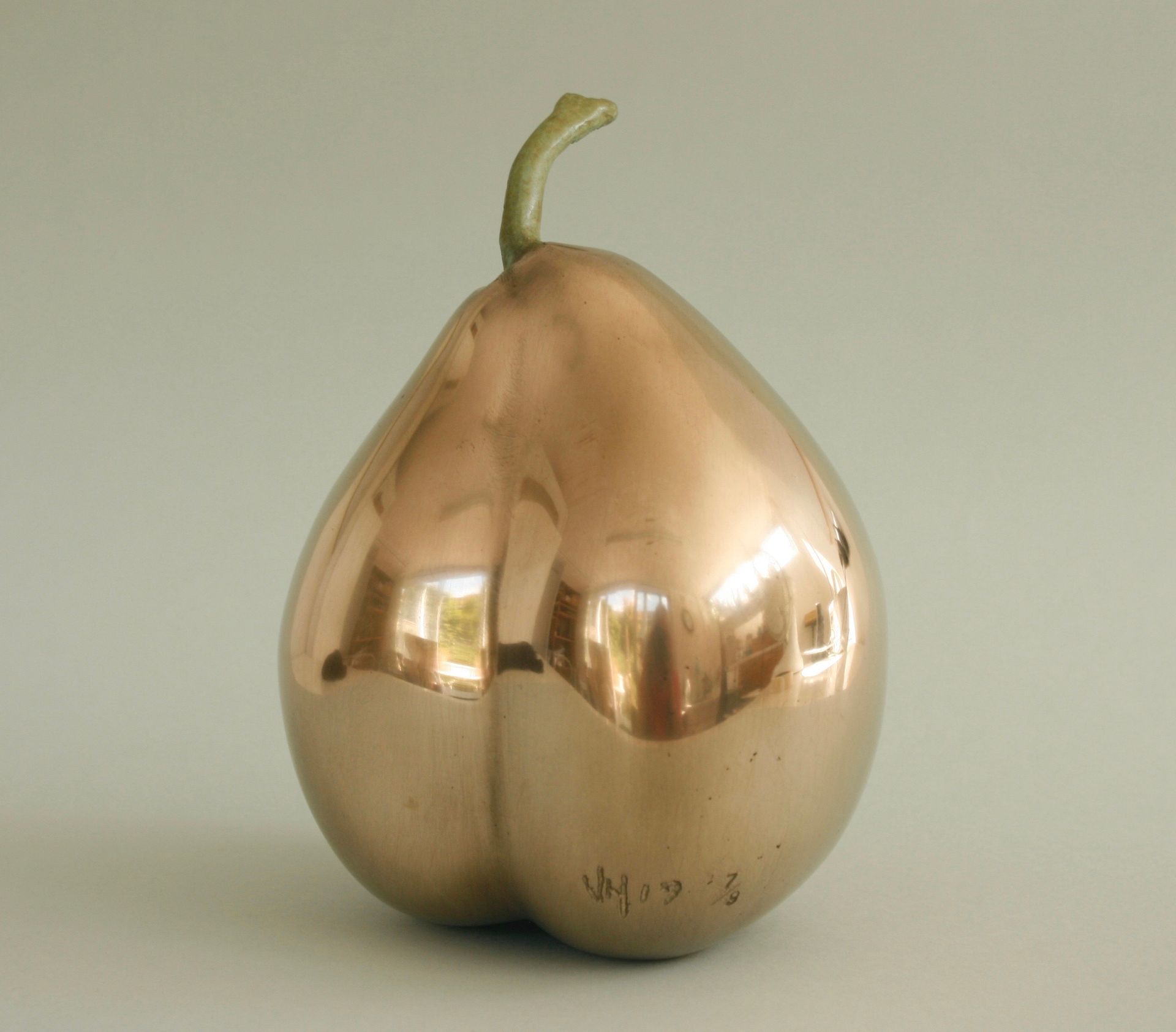 A polished copper pear sculpture with a green stem, reflecting its surroundings.