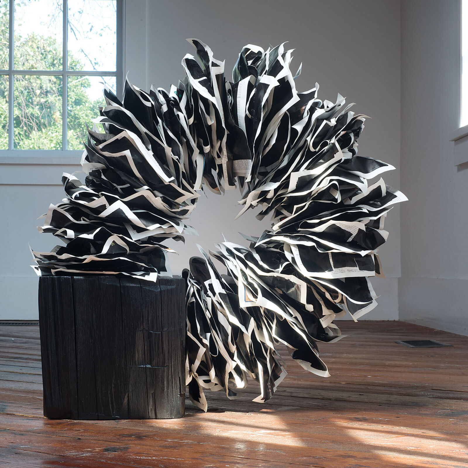 A black and white sculptural installation featuring a zebra-patterned wreath resting on a dark wooden stump.