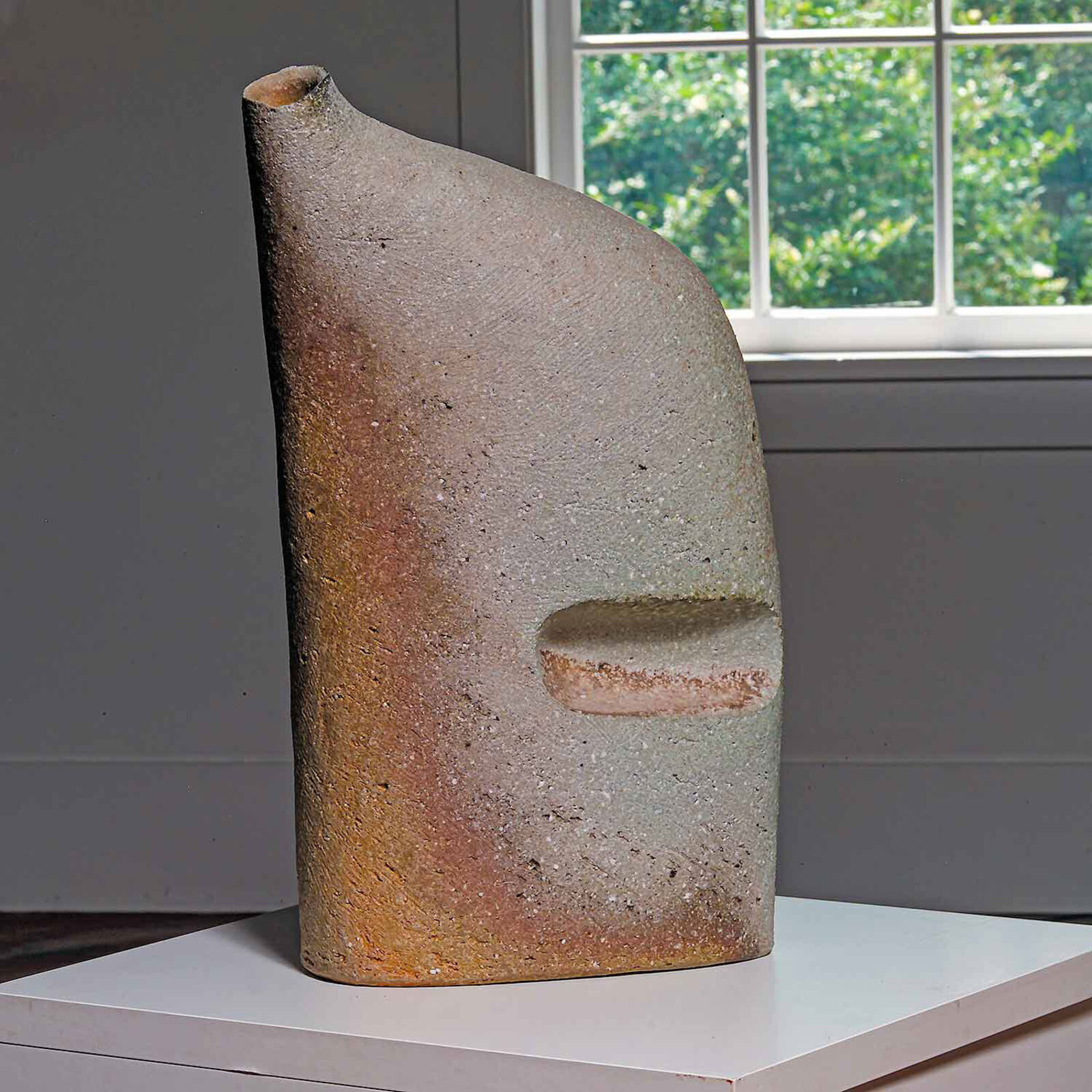 A textured, abstract sculpture with a face-like form and a recessed mouth sits on a white pedestal.