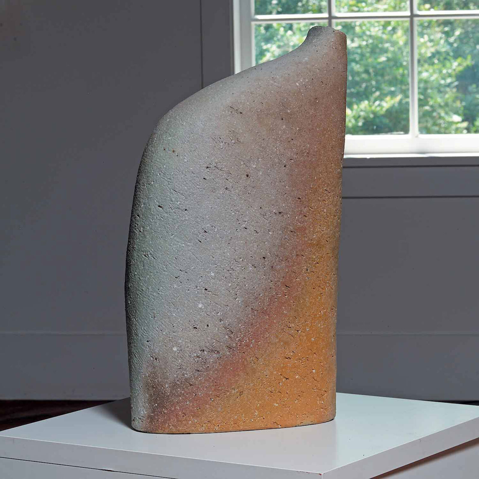 A textured, abstract sculpture with a face-like form and a recessed mouth sits on a white pedestal.