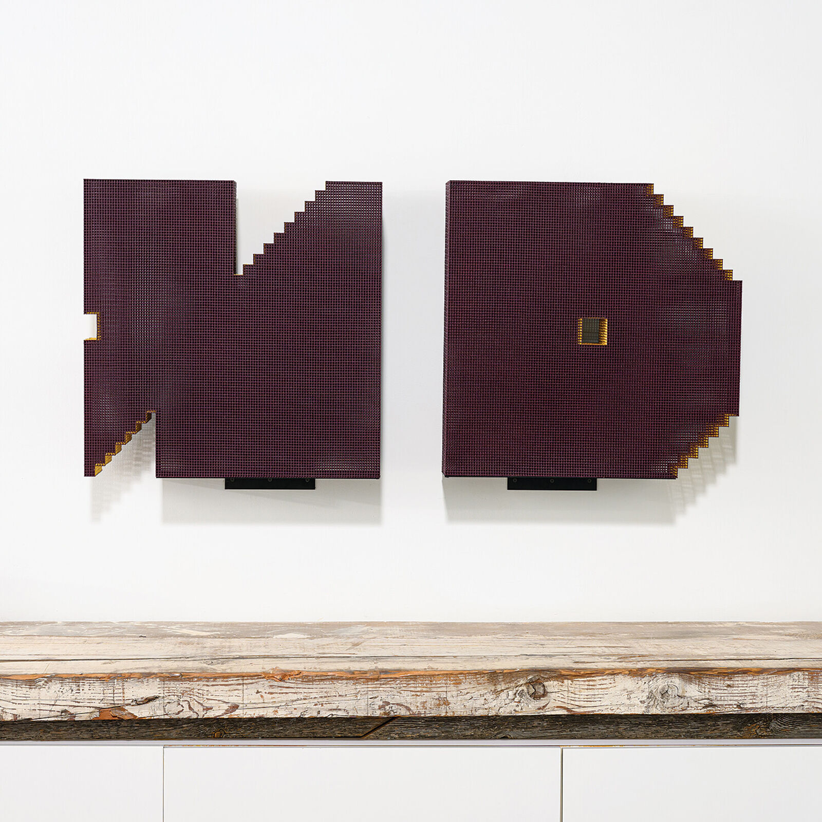 A minimalist installation featuring two pixelated, maroon-colored panels mounted on a white wall above a weathered wooden shelf.