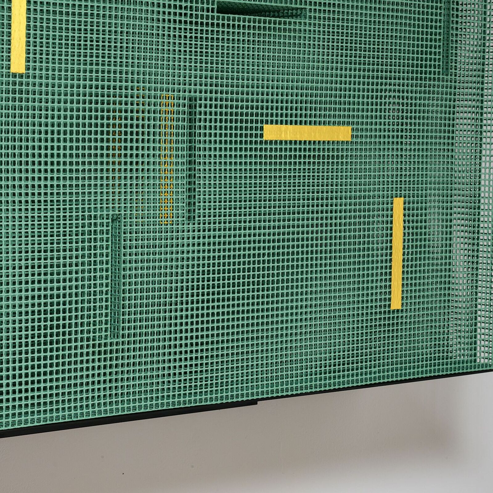 A green mesh sculpture with yellow and black geometric accents mounted on a white wall.
