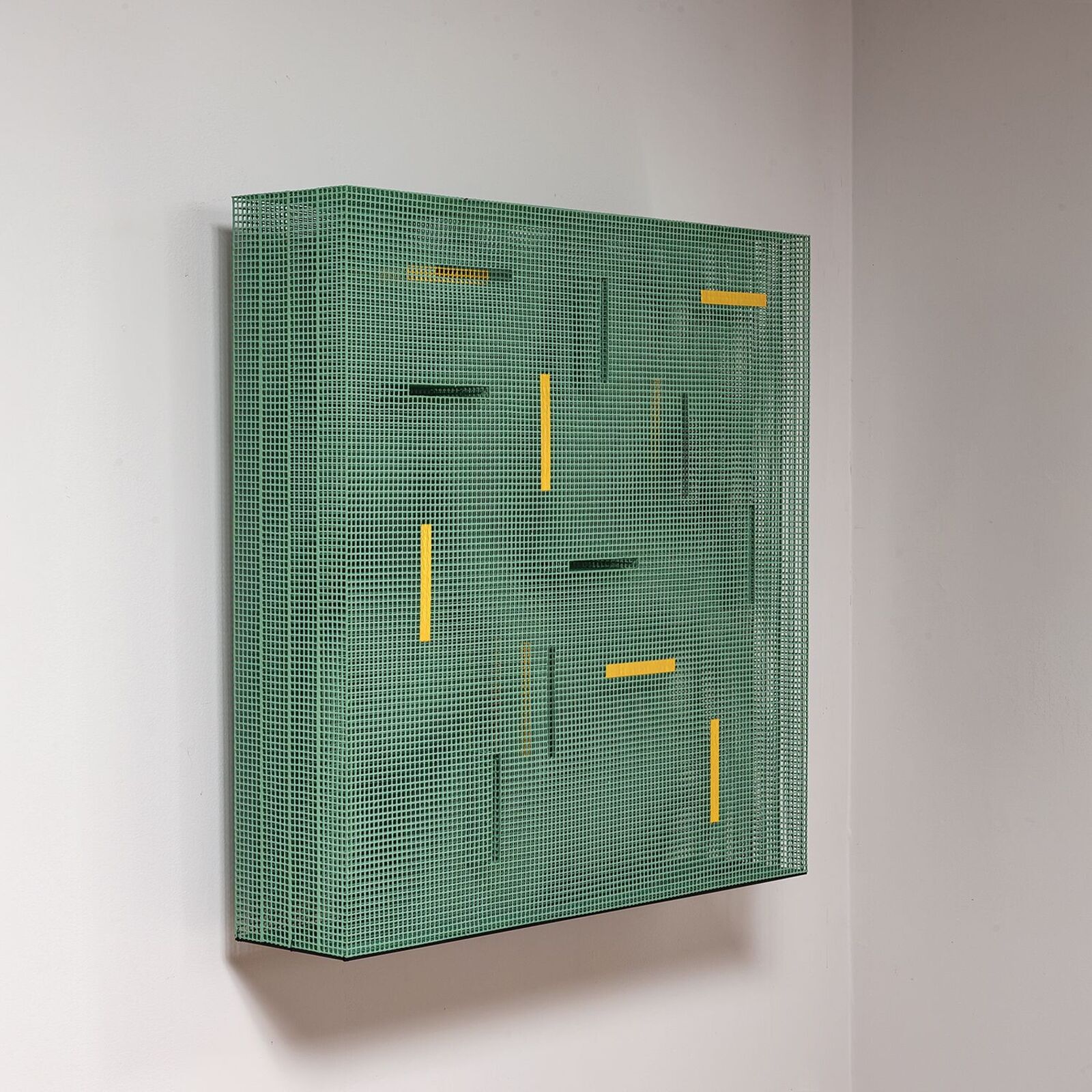 A green mesh sculpture with yellow and black geometric accents mounted on a white wall.