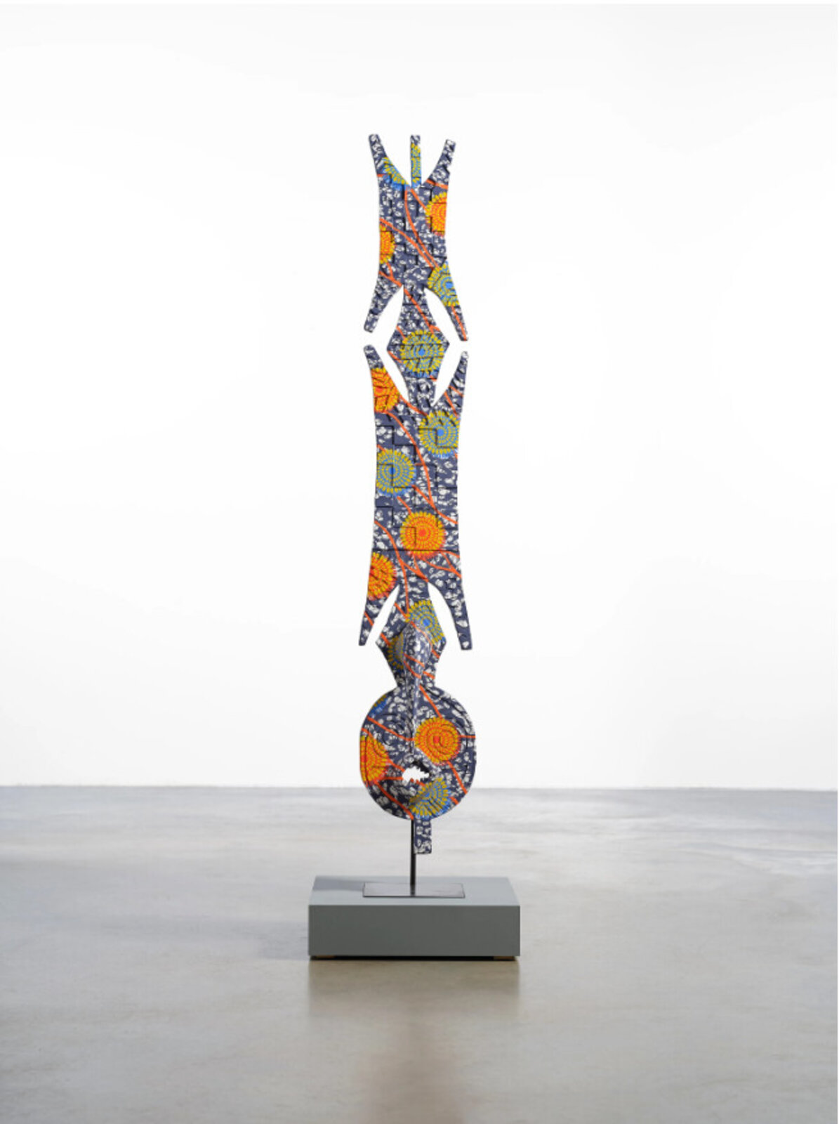 A tall, patterned sculpture with organic shapes and bold colors stands on a minimalist pedestal.