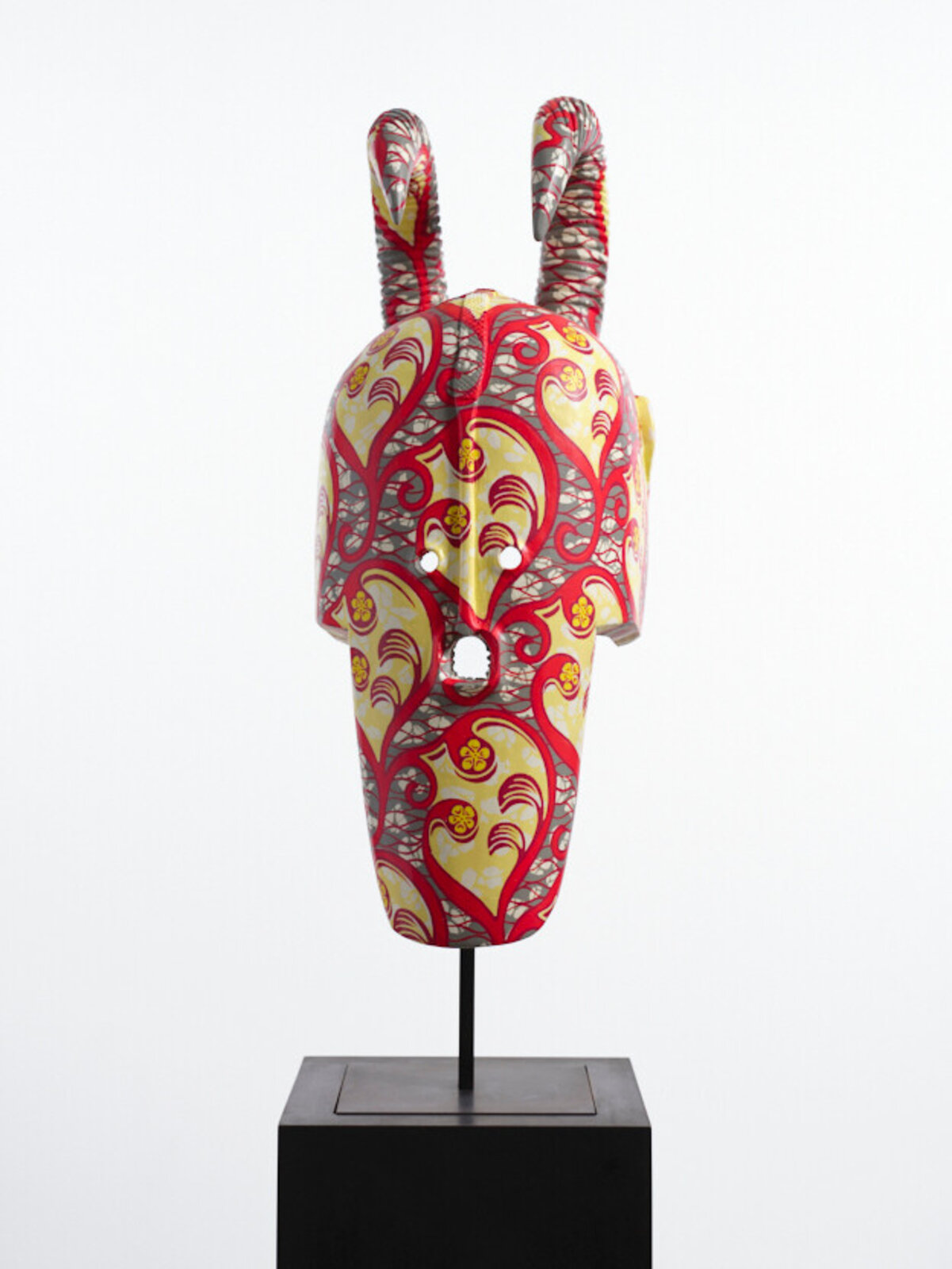 A colorful, patterned mask with curved horns displayed on a black pedestal.